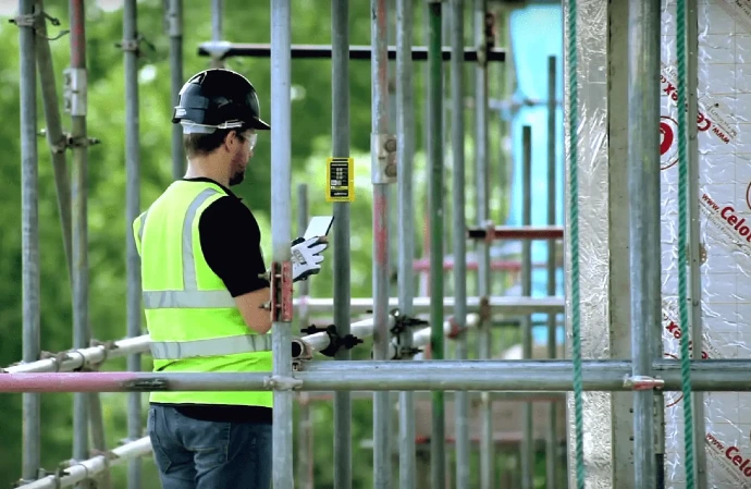 Construction worker wearing safety equipment on scaffolding, scanning an Inspect7® inspection device using a mobile device. 