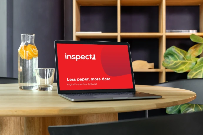 Laptop on a round wooden table displaying the Inspect7® logo on red background with the tagline ‘Less paper, more data’, set in a modern office space with shelving, a water carafe, and indoor plants.